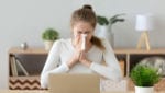 Woman sneezes into tissue while working on laptop