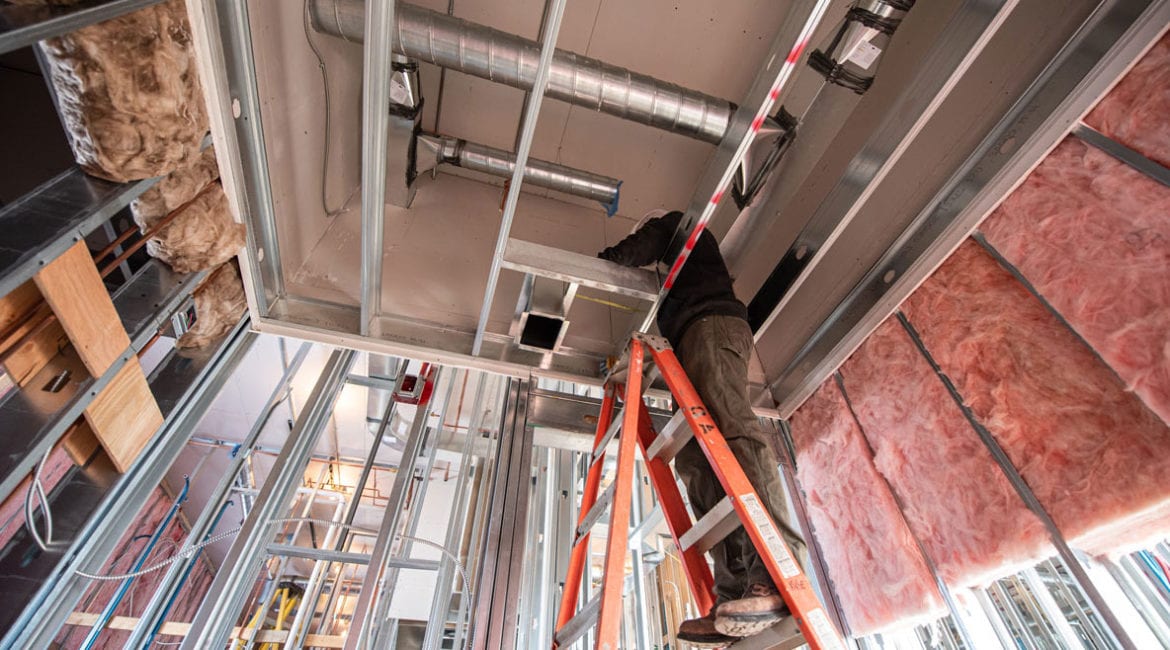 An HVAC professional stands on a ladder to install ductwork during the construction of a building