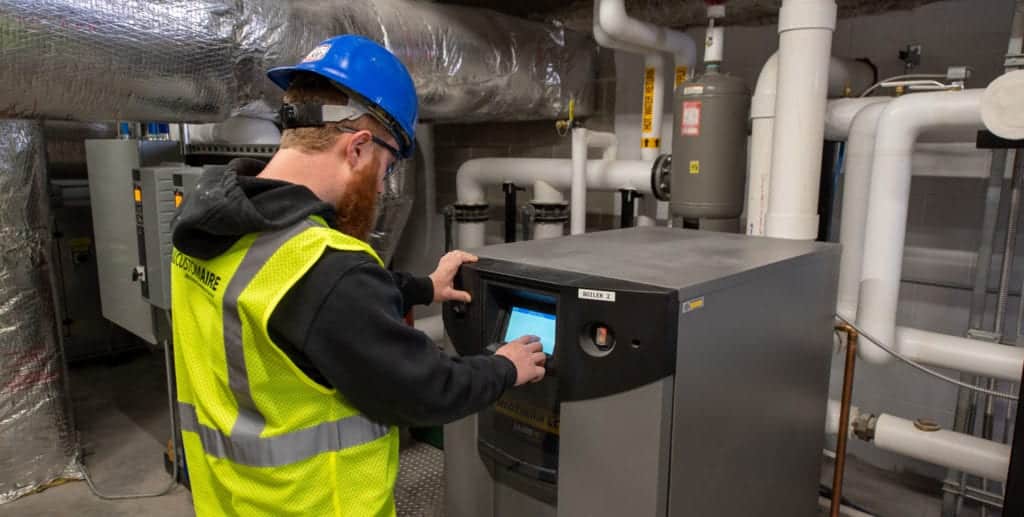 A male Custom Aire HVAC service technician inspects a LAARS commercial boiler in the mechanical room of an apartment building
