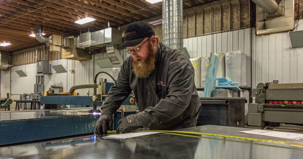 A male Custom Aire sheet metal fabrication technician measures a piece of sheet metal in the company’s fab shop
