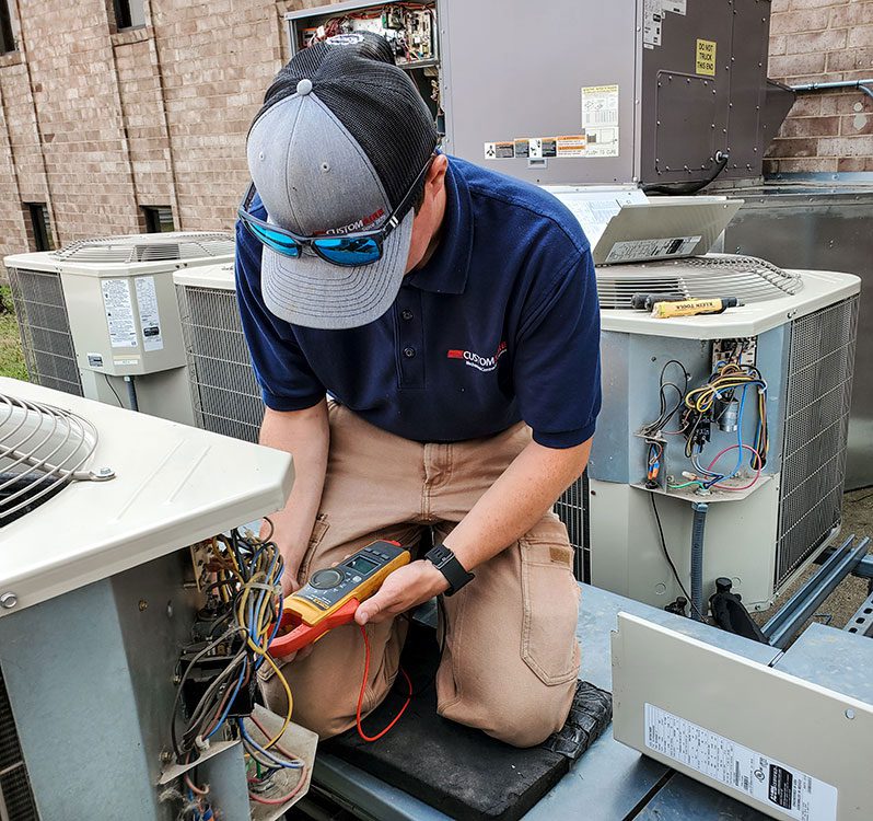 A male Custom Aire HVAC service technician performs routine maintenance of a commercial air conditioning system at a business